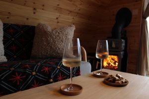 Shelter Pod interior coffee table with glasses of wine and lit wood-burning stove, North Yorkshire